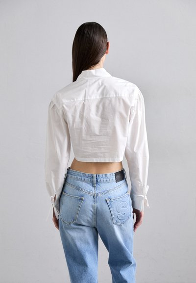 Avklippt vit blus med puffärmar, knutna manschetter, i kombination med ljusblå högmidjade jeans som har en sydd bokstav 'S' på fickan.