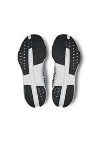 Semelles de chaussures de sport noir et blanc avec un caoutchouc texturé, dotées de trous de drainage, d'une rainure centrale et d'une section intermédiaire lisse blanche.