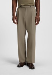 Man wearing straight-fit beige dress trousers and dark brown leather loafers, standing against a plain light background.