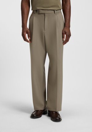 Man wearing straight-fit beige dress trousers and dark brown leather loafers, standing against a plain light background.