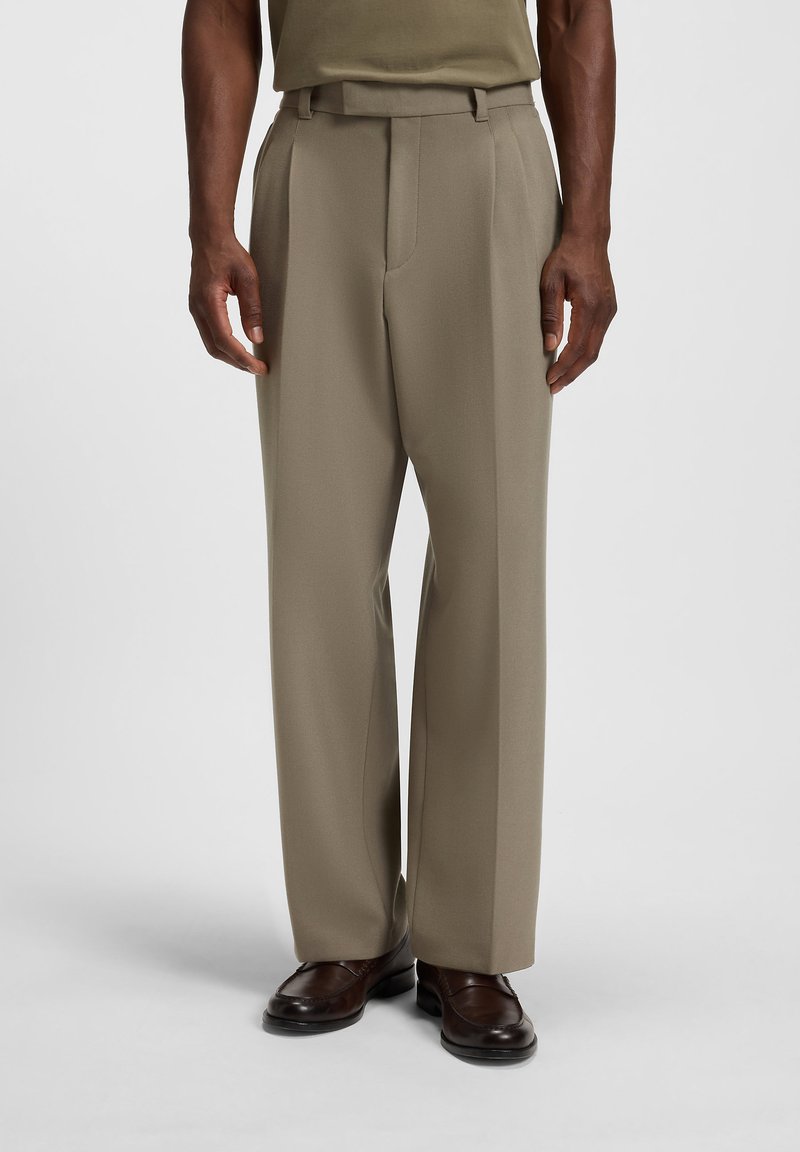 Man wearing straight-fit beige dress trousers and dark brown leather loafers, standing against a plain light background.