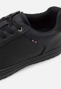 Crne tenisice s teksturiranim sintetičkim gornjištem, dizajnom s vezicama, srebrnim rupicama i malim višebojnom logom sa strane. Niska profil potplat.