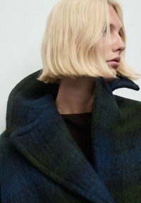Cappotto in misto lana in tonalità di verde scuro e blu, caratterizzato da un colletto alto, una forma strutturata e una sottile texture. Design semplice e moderno.
