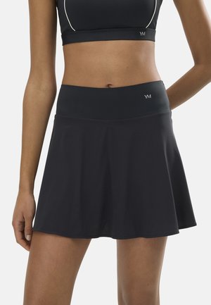 Mujer con una falda deportiva negra y sujetador deportivo a juego, de pie con una mano detrás de la espalda sobre un fondo claro.