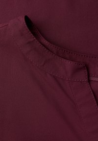 Bordeaux-stof med en glat overflade, prydet med en foldet krave og synlige syninger langs sømmen. Ingen mønstre eller hardwaredetaljer til stede.