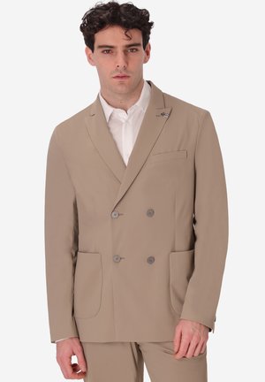 Uomo con capelli scuri e ricci che indossa un blazer beige doppiopetto sopra una camicia bianca, in piedi davanti a uno sfondo bianco neutro.