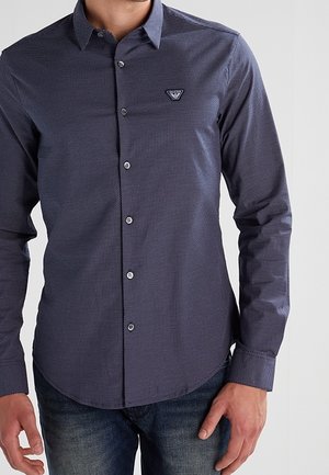 Homme portant une chemise à manches longues ajustée bleu foncé à motif avec un petit logo triangulaire sur la poitrine, associée à un jean bleu.