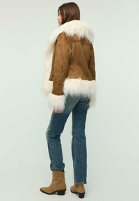 Brun ruskindjakke med hvid kunstpelskrave og manchetter, kombineret med blå denimjeans og lysebrune cowboystøvler. Ensfarvet baggrund.