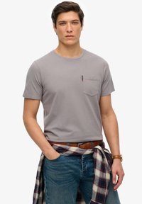 Grijze T-shirt met korte mouwen en een borstzak, gecombineerd met blauwe jeans. Een geruit shirt is om de taille geknoopt. Eenvoudig en functioneel ontwerp.