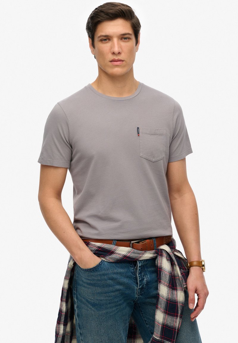 Grijze T-shirt met korte mouwen en een borstzak, gecombineerd met blauwe jeans. Een geruit shirt is om de taille geknoopt. Eenvoudig en functioneel ontwerp.