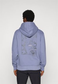 Mand iført en lavendelfarvet hoodie med et sort og gråt grafisk design af en skiløber, der kører ned ad et bjerg på ryggen, parret med sorte bukser.