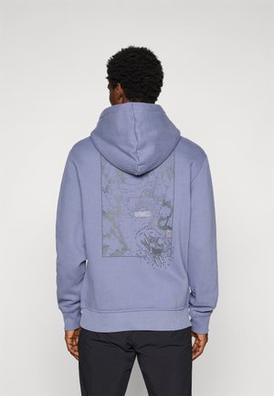 Mann trägt einen lavendelfarbenen Hoodie mit einem schwarz-grauen Grafikmotiv eines Skifahrers, der einen Berg hinunterfährt, auf dem Rücken, kombiniert mit schwarzen Hosen.