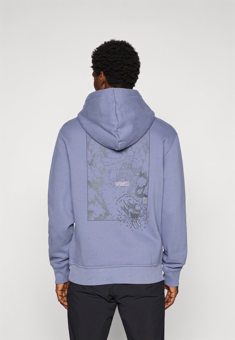 Mand iført en lavendelfarvet hoodie med et sort og gråt grafisk design af en skiløber, der kører ned ad et bjerg på ryggen, parret med sorte bukser.