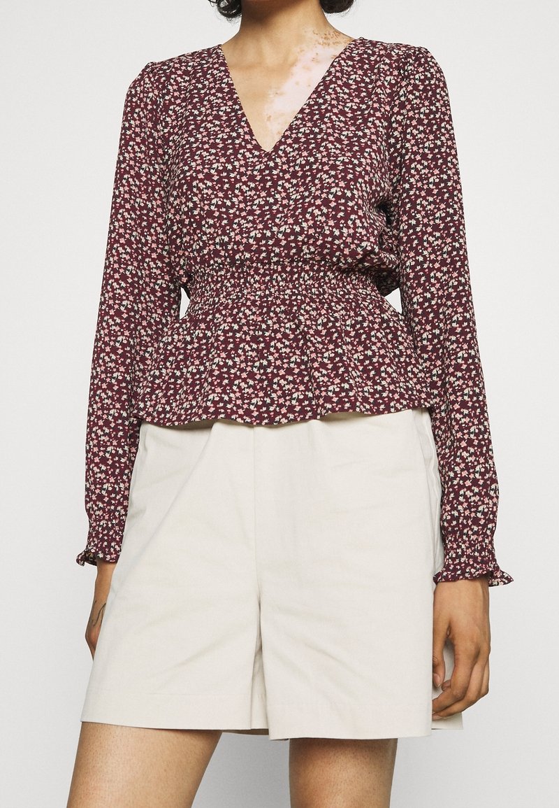 Femme portant un blouse à manches longues bordeaux à motifs floraux avec une taille smockée et un short taille haute beige sur un fond uni.