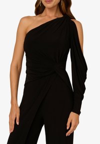 Top noir à une épaule avec du tissu drapé, détail noué à la taille, manche longue d'un côté et texture lisse.