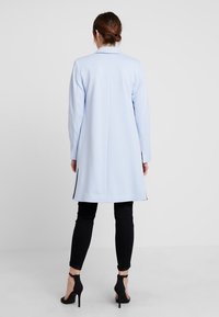 Donna con i capelli castani raccolti in un bun, indossa un soprabito azzurro chiaro fino al ginocchio, pantaloni skinny neri e scarpe col tacco nere, in piedi contro uno sfondo neutro.
