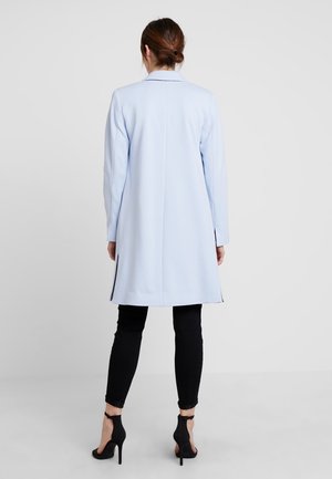Donna con i capelli castani raccolti in un bun, indossa un soprabito azzurro chiaro fino al ginocchio, pantaloni skinny neri e scarpe col tacco nere, in piedi contro uno sfondo neutro.