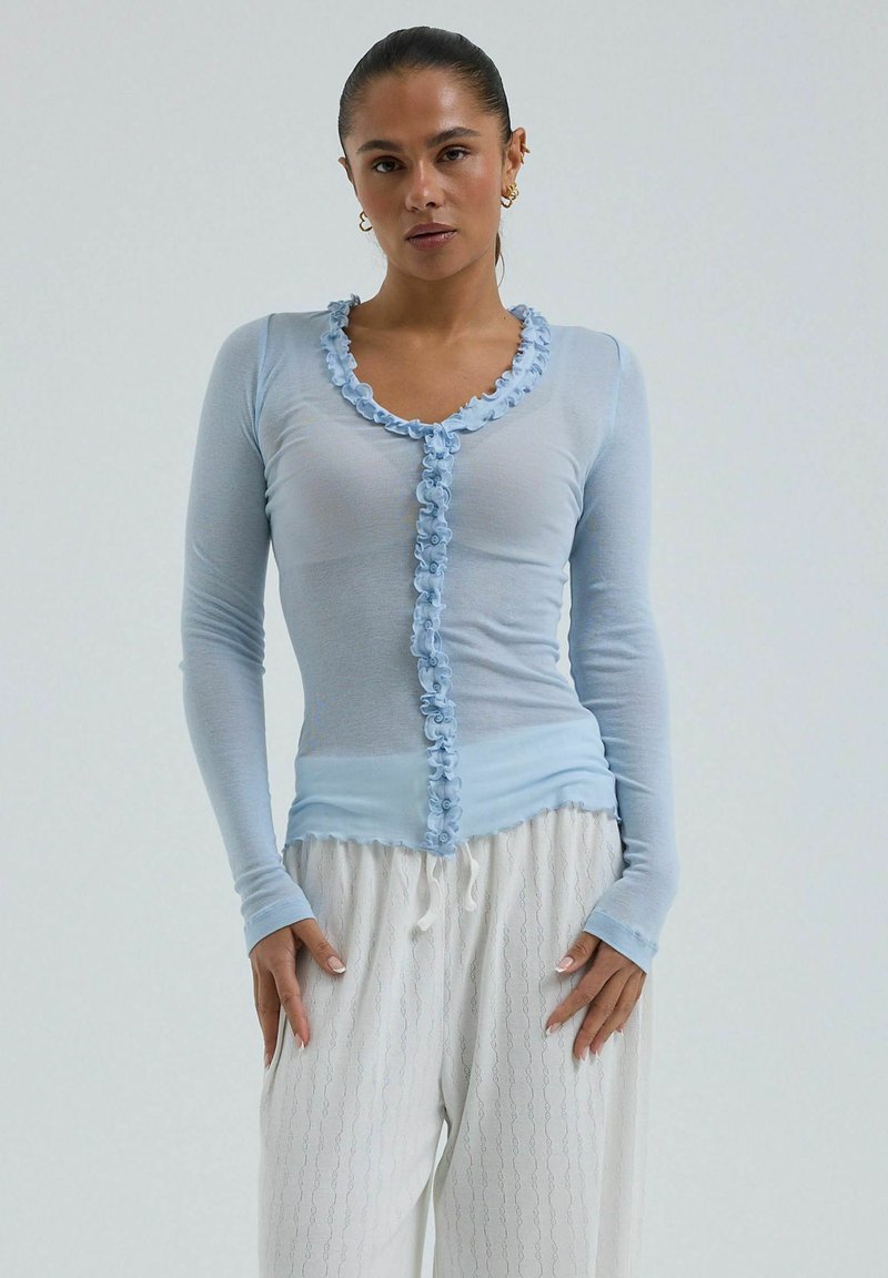 Femme portant un haut bleu clair à manches longues avec un bord à volants et un pantalon blanc ample avec un motif subtil, debout sur un fond uni.