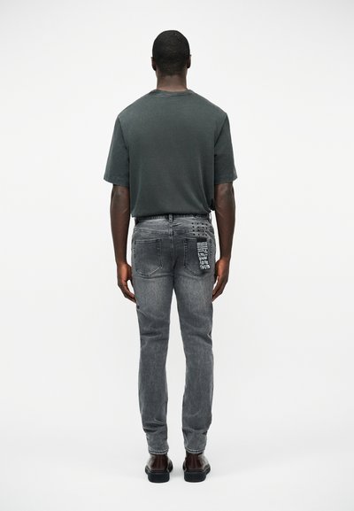 Man staat met zijn rug naar de camera, draagt een donkergrijs shirt met korte mouwen en vervaagde zwarte jeans met witte tekst en ontwerp op de rechterachterzak.