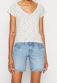 Top blanco de manga corta con un patrón floral y bordes ondulados, combinado con unos pantalones cortos de mezclilla azul. El conjunto presenta detalles de costura simples.