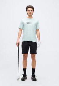 Νέος άνδρας που στέκεται με μια ράβδο γκολφ, φορώντας ανοιχτόμπλε μπλουζάκι, μαύρο σορτς, μαύρες κάλτσες, μαύρα παπούτσια και ένα λευκό γάντι γκολφ.