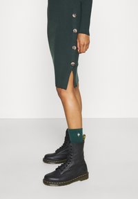 Mustat nahkakombat-kengät, joissa on keltaiset tikkaukset ja läpinäkyvät pohjat. Mallilla on päällään vihreä ribbimekko, jossa on polven korkeudet halkiot ja messinkiset napit.