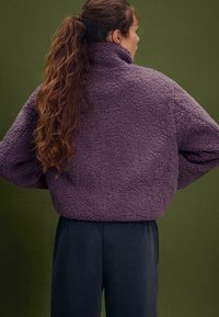 Giacca in pile viola e morbida con colletto alto e spalle abbassate. Indossata sopra pantaloni scuri, con una superficie texturizzata.