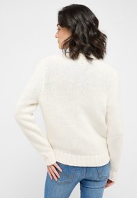 Stickad krämfärgad tröja med ribbad nederkant och ärmslut, med en lös passform och öppen vävstruktur, stylad med blå jeans.