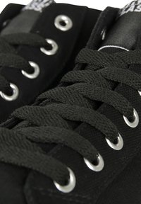 Mustad mustadest kanva sneakerid, metallist silmade ja paksude mustade paeltega. Ülemine osa on tekstuuriga viimistlusega, rõhutades õmblusi ja metallosi.