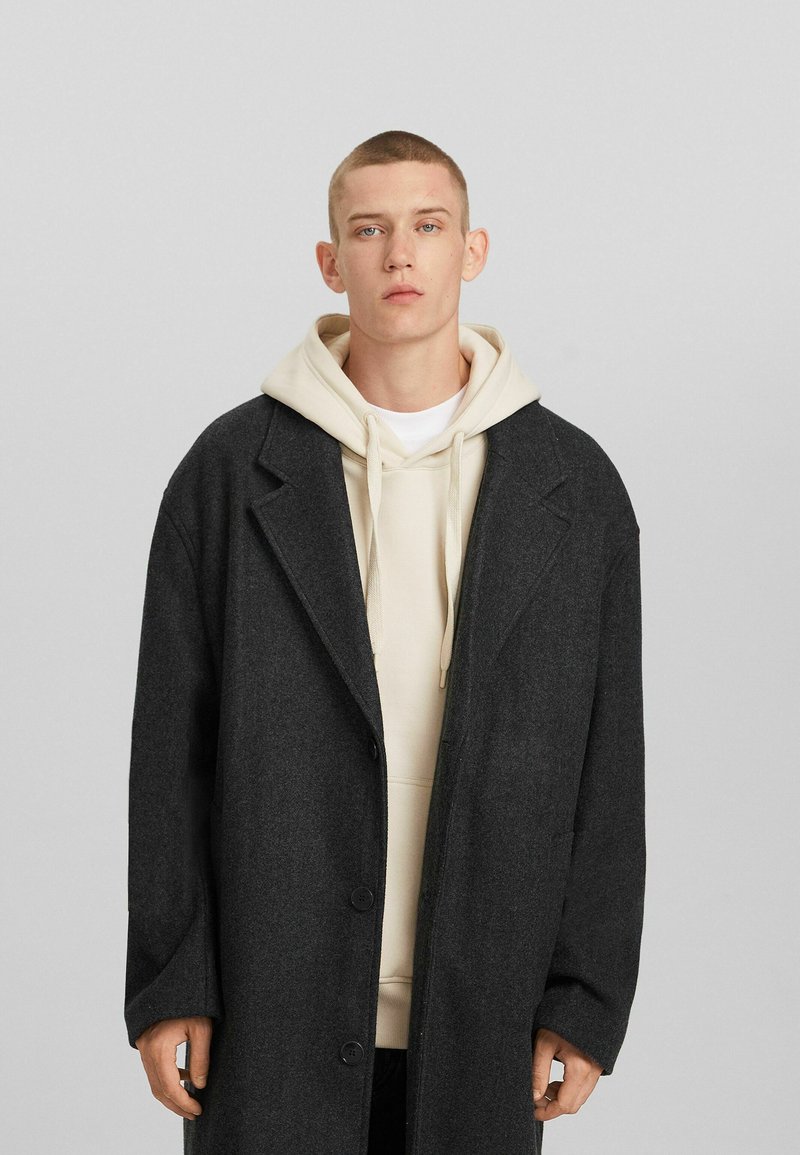 Jonge man in een donkergrijze overjas over een beige hoodie, staand tegen een effen lichtgrijze achtergrond, recht vooruit kijkend.