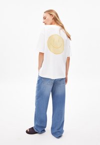 Eine Frau mit langen blonden Haaren steht in einem lässigen weißen T-Shirt mit einem großen gelben Smiley auf dem Rücken, blauen weiten Jeans und schwarzen Schlappen.