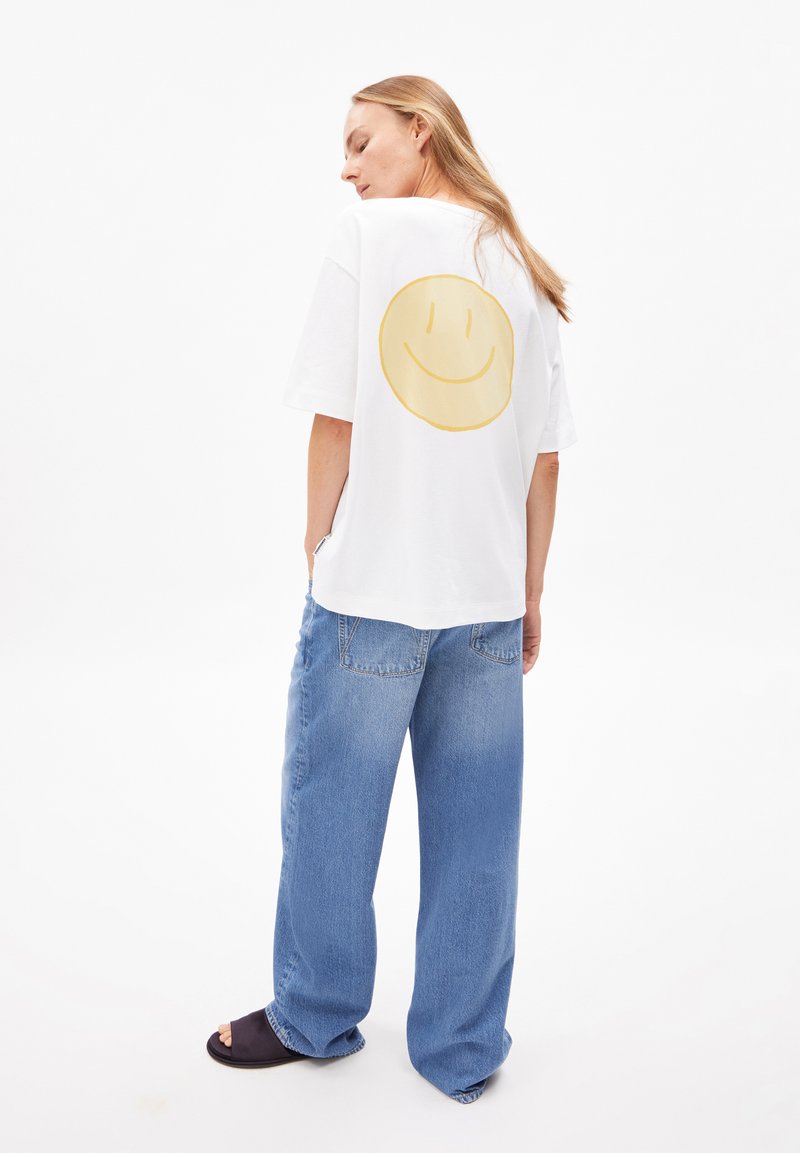 Eine Frau mit langen blonden Haaren steht in einem lässigen weißen T-Shirt mit einem großen gelben Smiley auf dem Rücken, blauen weiten Jeans und schwarzen Schlappen.