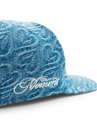 Denimkeps med en texturerad blå tyg som har ett upprepande mönster, och en vit broderad "Moment"-logotyp på framsidan.