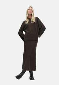 Ruskea neulotun villatakin, jossa on ylisuuressa hihat, yhdistettynä samanlaiseen pitkään hameeseen. Mallilla on mustat nilkkurit, joissa on sileä pinta.