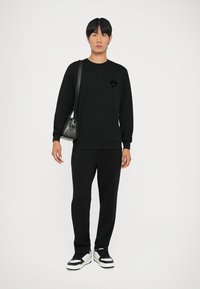 Felpa nera con un'emblema testurizzato, abbinata a pantaloni neri e sneaker neri con dettagli bianchi, insieme a una piccola borsa a tracolla nera.