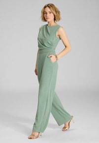 Combinaison de couleur vert menthe avec un décolleté drapé et une conception à jambes larges. Elle possède des poches latérales et une texture lisse. Des sandales à talons dorées rehaussent le look.