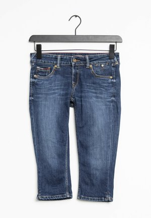 Blaue, verkürzte Jeans mit vorderen Taschen und Gürtelschlaufen, die an einem schwarzen Plastikbügel vor weißem Hintergrund hängen.