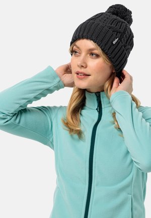 Berretto lavorato a maglia grigio scuro con texture a coste e pompon in cima, indossato con una giacca in fleece azzurro chiaro.
