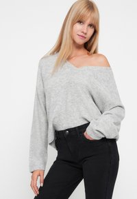 Szary, oversize sweter z szerokim dekoltem w kształcie litery V i wykorzystującym design bez rękawów. Miękka tekstura, prążkowane mankiety, w zestawieniu z czarnymi spodniami z wysokim stanem.
