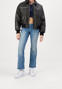 Cazadora bomber de piel negra con puños de canalé, bolsillos con cremallera y un diseño corto, combinada con vaqueros acampanados de color azul claro y zapatillas blancas.