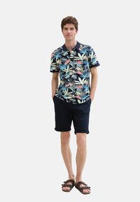 Herre kortærmet polo med et flerfarvet tropisk bladmønster, marinefarvet krave og ærmer, sat sammen med marinefarvede shorts og brune sandaler.