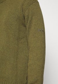 Närbild av en olivgrön stickad tröjsleeve med subtilt broderad "BOSS"-logotyp nära sömmen på överarmen.