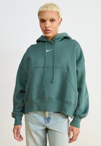 Άτομο με κοντά ξανθά μαλλιά, φορώντας μια διαμαντέ μπλούζα Nike και ανοιχτόχρωμα μπλε τζιν, στέκεται μπροστά σε μια λευκή, απλή επιφάνεια.