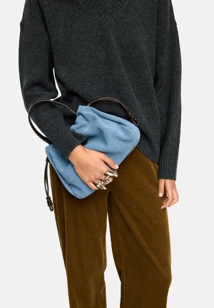 Osoba ubrana w ciemnoszary sweter i brązowe spodnie sztruksowe trzymająca miękką niebieską torebkę z ciemnym uchwytem i nosząca duże srebrne pierścionki.