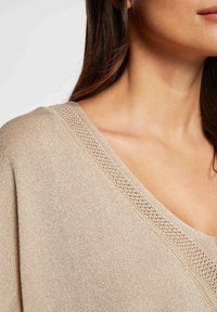 Beige gebreide trui met een brede V-hals en ingewikkelde open gebreide details bij de halslijn, met een zachte, gestructureerde afwerking.