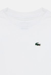 Λευκό μπλουζάκι με στρογγυλή λαιμόκοψη και μικρό πράσινο λογότυπο κροκόδειλου στο αριστερό στήθος, εμφανίζοντας λεπτομέρειες στον γιακά και υφή υφάσματος.