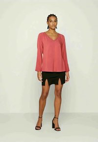 Top de manga comprida, com decote em V, em tecido rosa, combinado com uma saia mini preta com aberturas laterais, colocado sobre um fundo neutro.