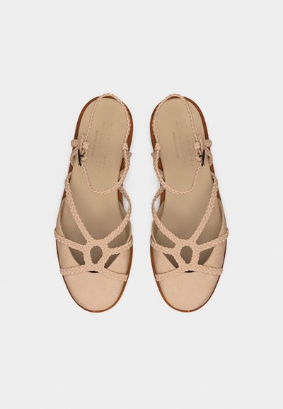 Ζευγάρι μπεζ επίπεδων σανδαλιών με πλεγμένους ιμάντες και αγκράφες στον αστράγαλο, εμφανιζόμενα από πάνω σε λευκό φόντο.