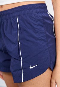 Jūrų mėlynos spalvos sportiniai šortai, pagaminti iš lengvos medžiagos, su baltu šoniniu apvadu ir mažu baltu „Nike“ logotipu kairiajame apatiniame kampe. Elastinė juosmens juosta.