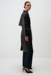 Cappotto in pelle nera con un design su misura e maniche corte. Presenta una cintura in vita, spacchi laterali ed è abbinato a jeans scuri e tacchi a punta.